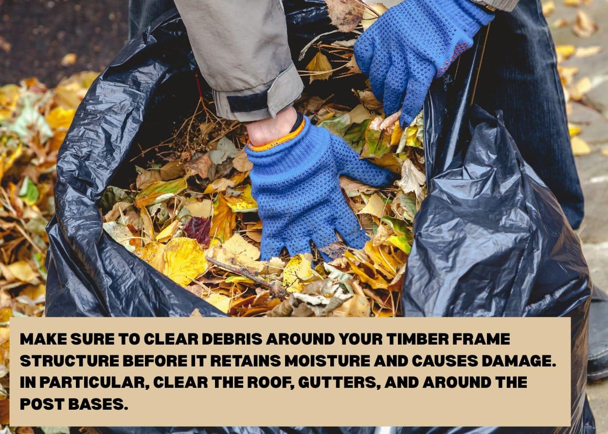Hands wearing work gloves collecting fallen leaves into a trash bag during autumn cleanup. Caption: Make sure to clear debris around your timber frame structure before it retains moisture and causes damage. In particular, clear the roof, gutters, and around the post bases.
