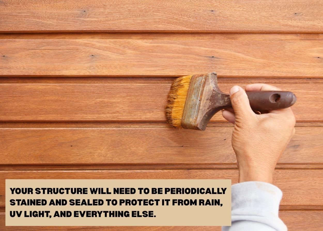 Person applying protective wood stain to a timber wall using a large brush for seasonal maintenance. Caption: Your structure will need to be periodically stained and sealed to protect it from rain, UV light, and everything else.